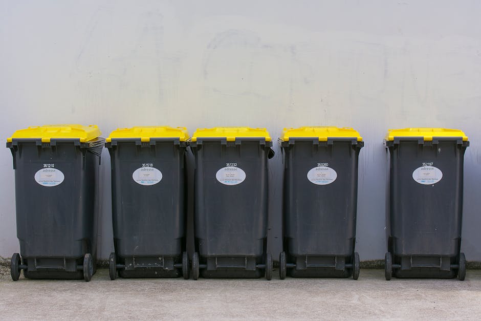 A green, old-fashioned clothing collection bin with a rectangular body and a sloped top, situated outdoors against a background of dense greenery and a wooden fence. The bin has compartments with smal