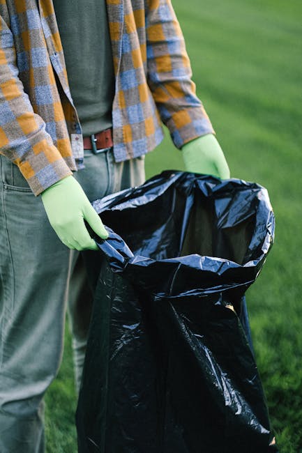 The image shows a person wearing a yellow and grey checkered shirt, grey trousers, and bright green gloves, standing outdoors on a grassy area. They are holding open a large black plastic rubbish bag,