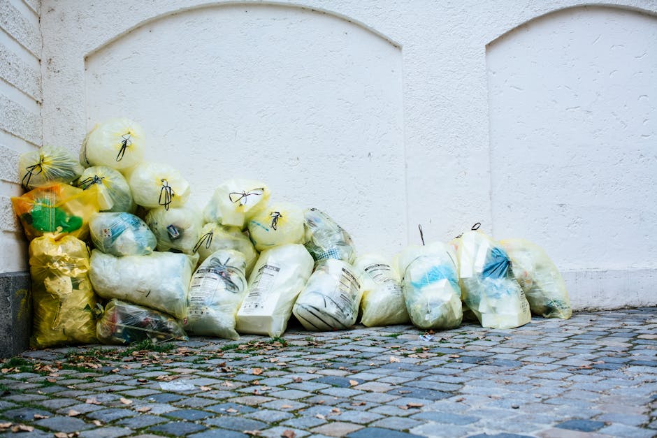The image shows a person wearing a yellow and grey checkered shirt, grey trousers, and bright green gloves, standing outdoors on a grassy area. They are holding open a large black plastic rubbish bag,